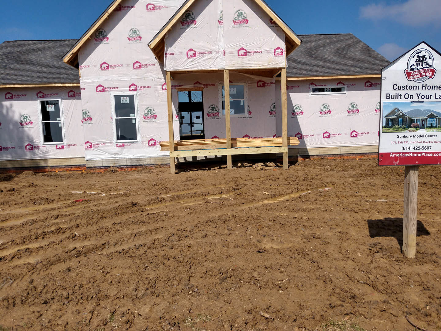 Two-story house under construction with exposed framing, plastic sheeting, and white-framed windows; dirt field in foreground with scattered wooden beams and a bench; cloudy sky