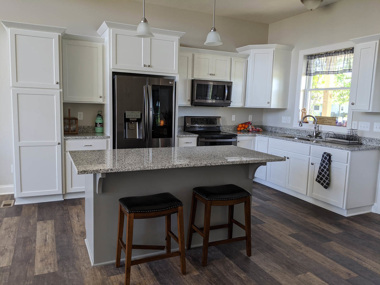 Open-concept kitchen featuring a bar with two black-cushioned stools, stainless steel refrigerator, built-in microwave, white cabinetry, stone countertops, and a window with a