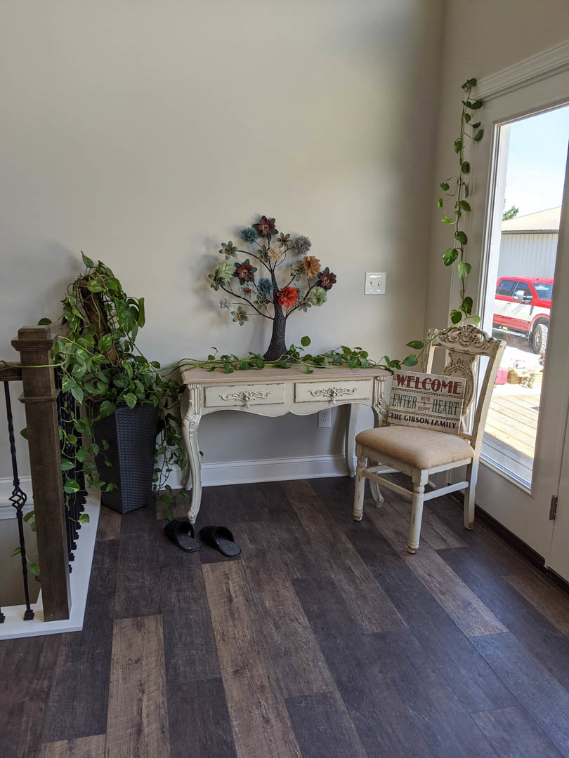 White table with a chair and potted plant on wood laminate flooring, black slippers nearby, neutral walls, and floral arrangement in a vase.