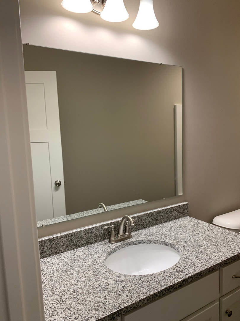 White bathroom with black speckled countertop, rectangular mirror above sink, chrome faucet, modern light fixture, and partial view of toilet against tiled wall
