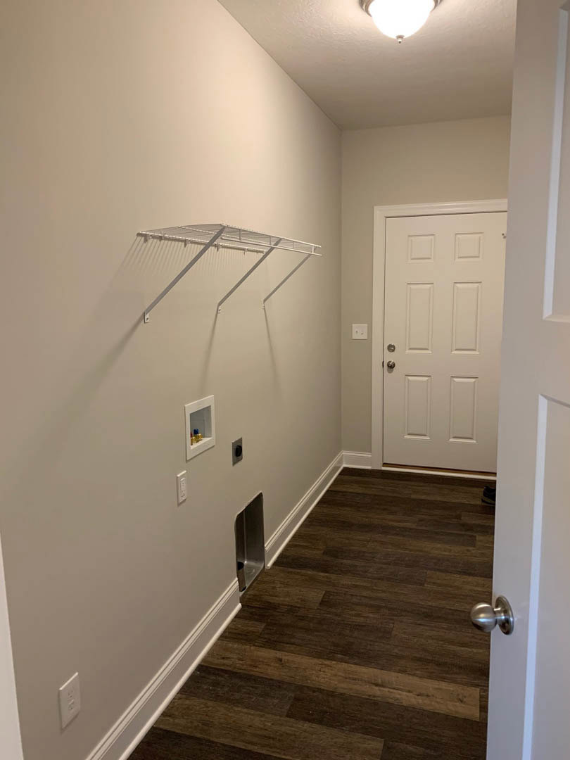 Hallway with light wood flooring, white door, white wall rack, ceiling light fixture, and a white shelf holding a small bottle