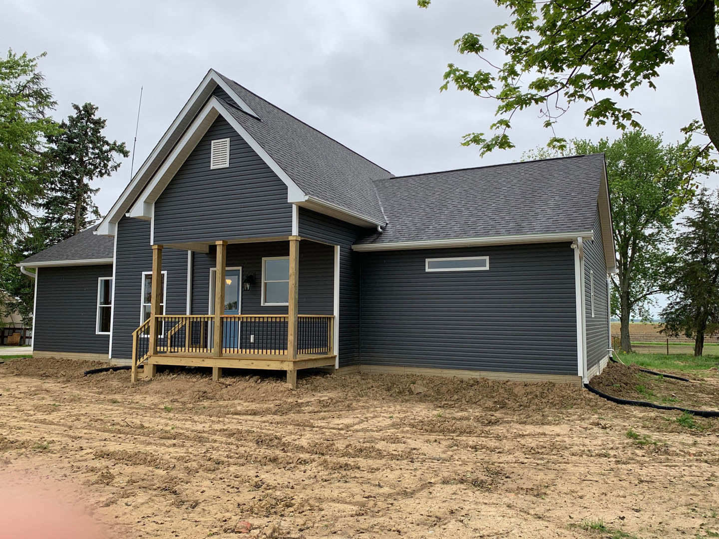 Wooden porch with white railing attached to a house featuring light siding, dirt path leading to entrance, tree branches with green leaves overhead, white-framed window visible.