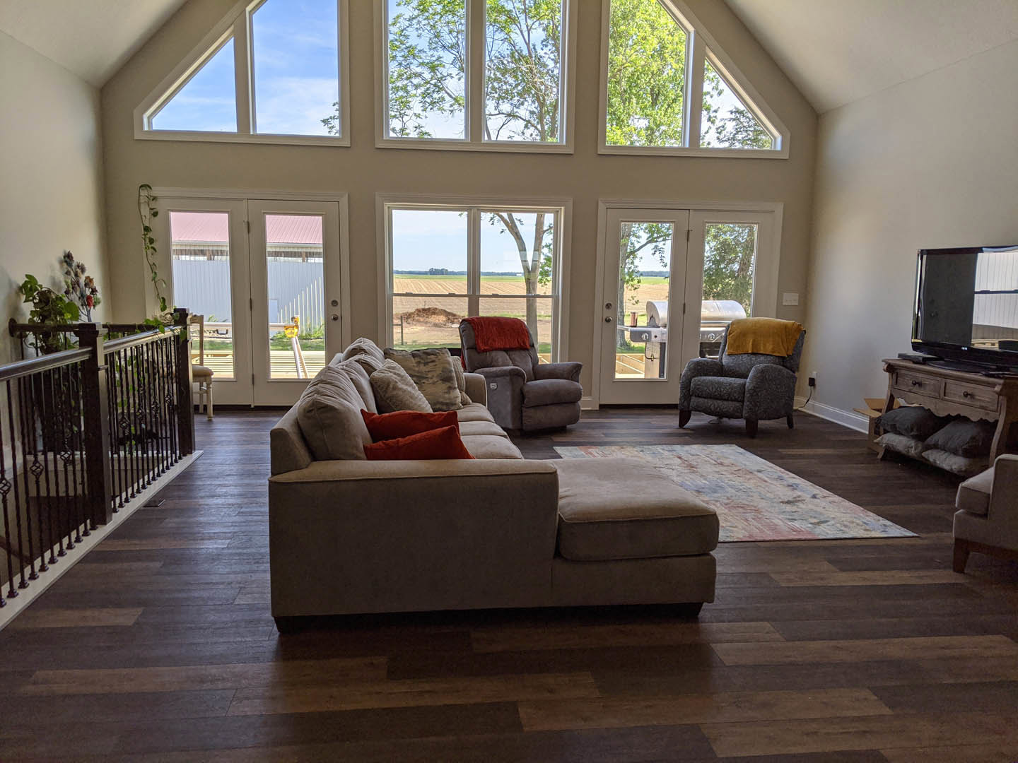 Living room with wide window, blue accent chair draped with yellow blanket, neutral sofa, staircase with wood railing, decorative pillow, wall-mounted television
