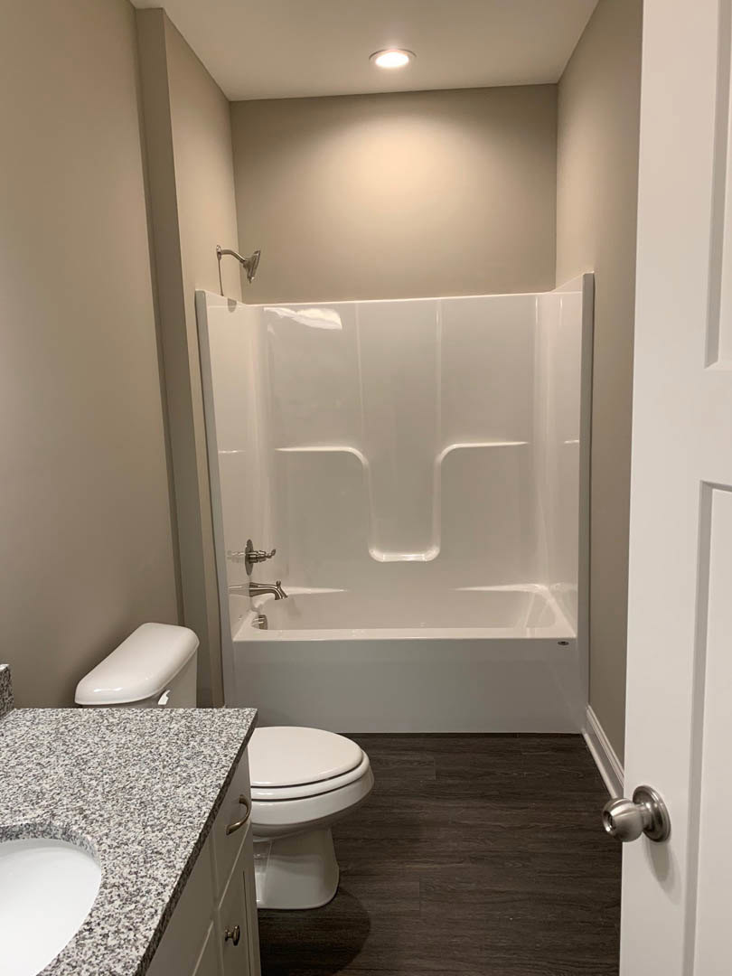 White bathtub with silver faucets, adjacent white toilet with matching tank and lid, light-colored tile walls, chrome bathroom fixtures, and a close-up of a modern sink and door