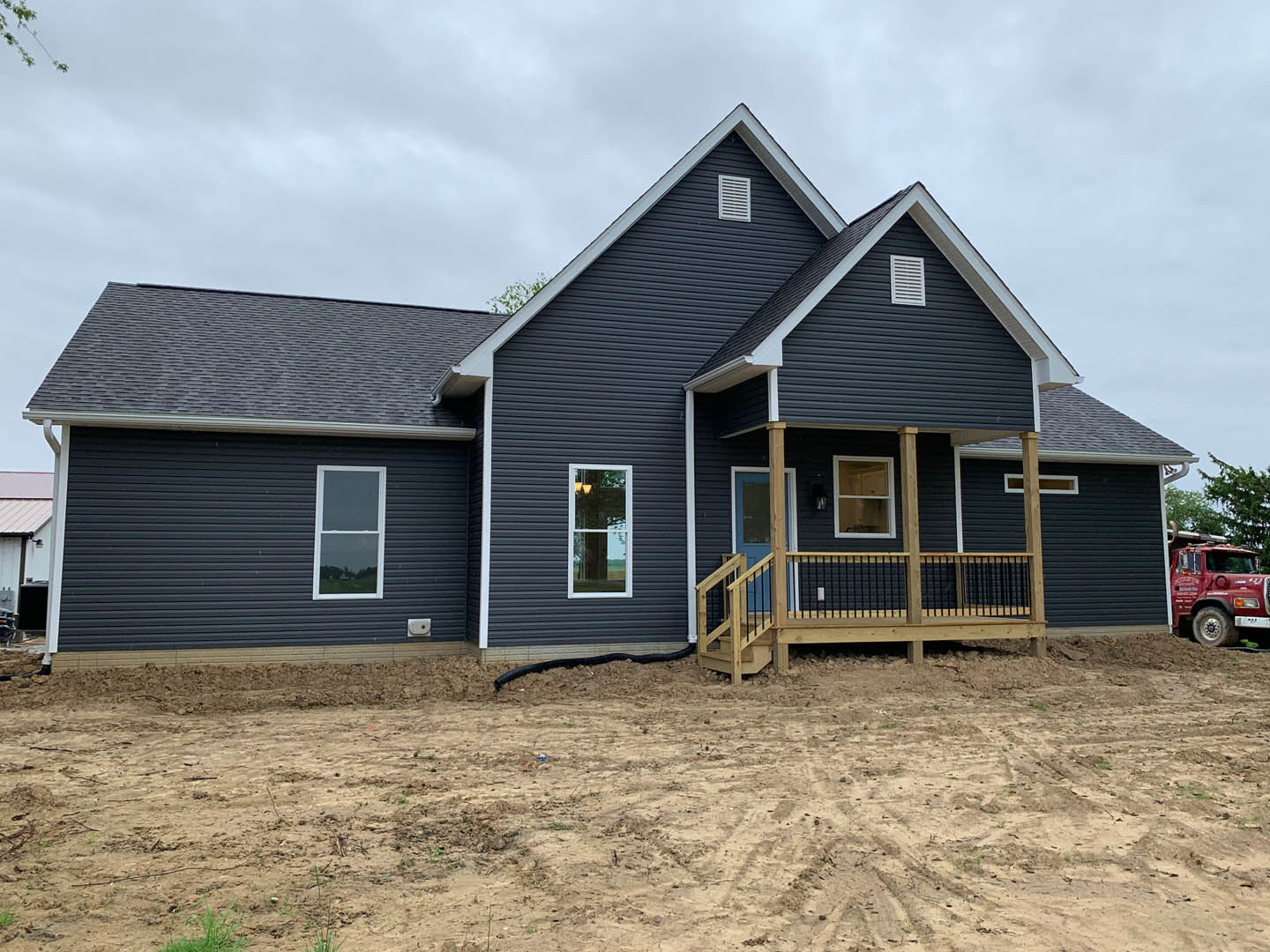 Two-story house with light siding, wooden deck and black railing, dirt yard in front, red truck parked roadside, porch with white-framed windows, cloudy sky overhead
