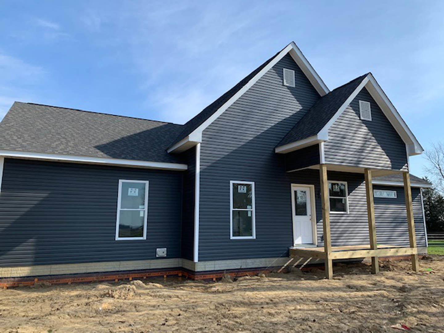 Wood-framed house under construction with covered porch, white framed windows, white door with glass panel, exposed siding, and construction signage visible on window