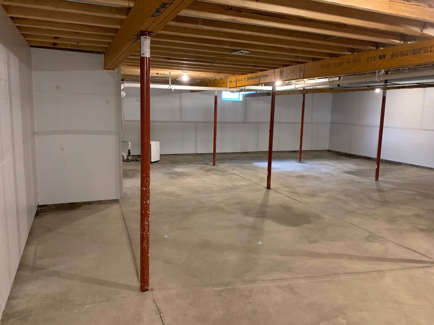 Open room with exposed wooden ceiling beams, white plaster wall featuring circular holes, concrete floor with red support poles, ceiling vent, red pole holding a white electrical