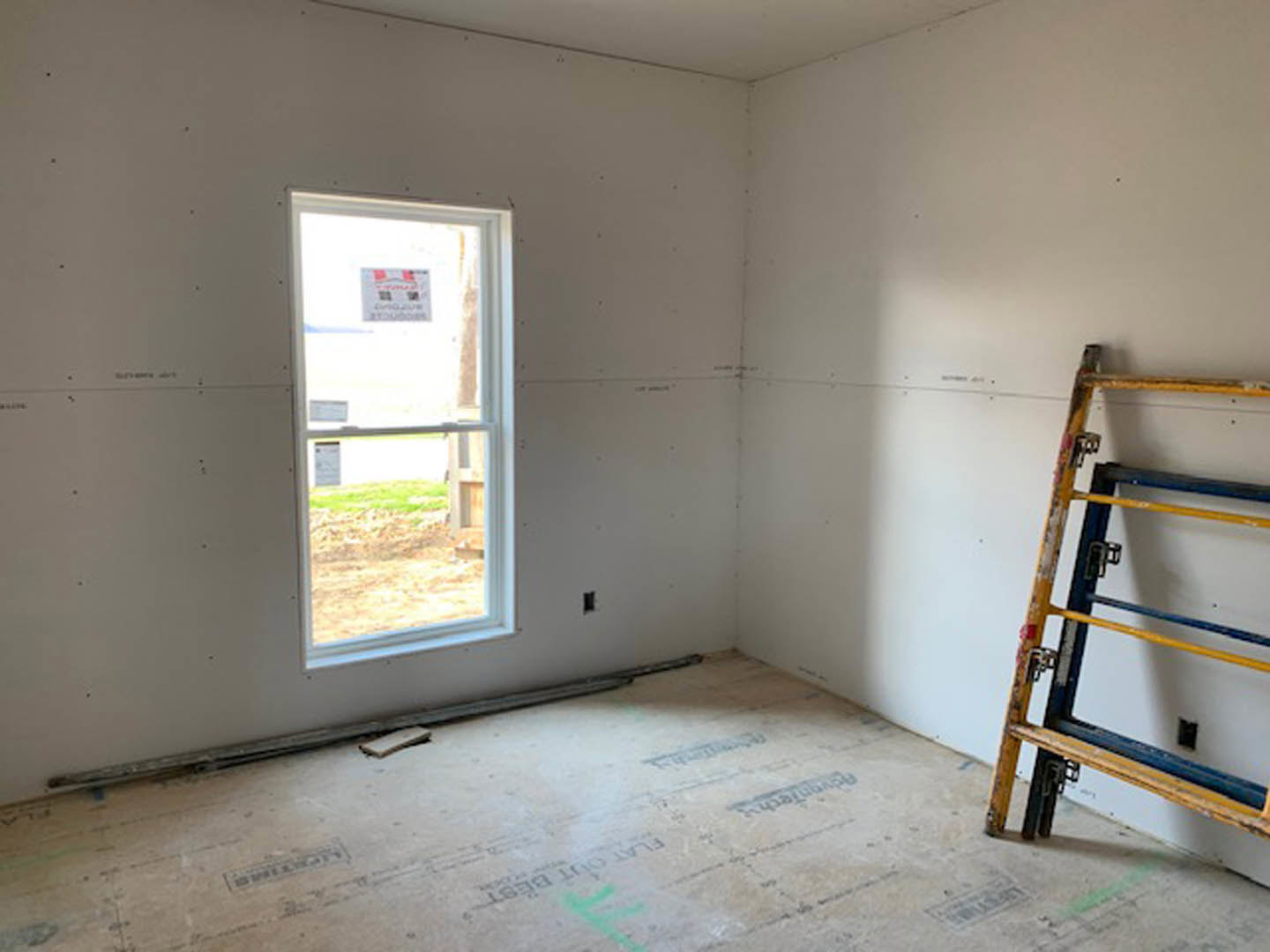 Ladder with yellow and blue bars standing on unfinished floor, sign taped to window, plastered walls, small piece of wood on floor, room under construction.