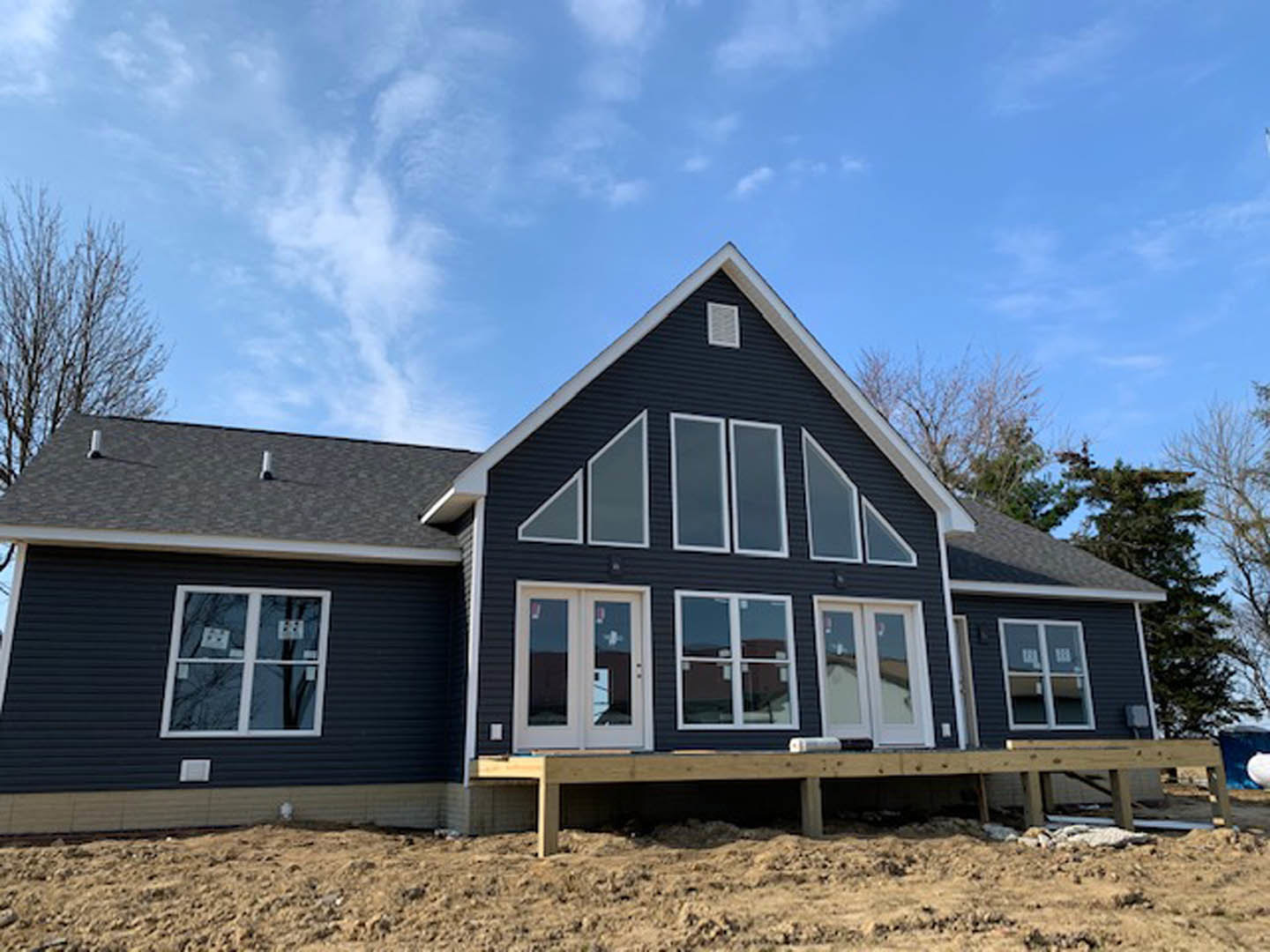 Framed custom home under construction with exposed wood, white-framed windows, double entry doors, black wall vent, dirt yard, and clear blue sky