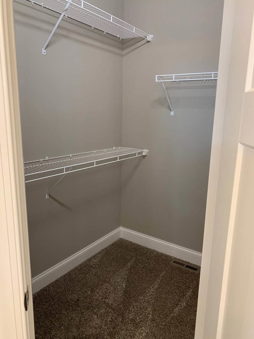 Walk-in closet with white shelving units featuring metal rails, beige carpet flooring, and a wall vent.