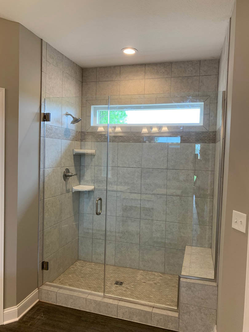 Glass shower enclosure with large window, white tile walls, recessed ceiling light, wood and tile flooring, white door, dual light switch, and modern plumbing fixtures.