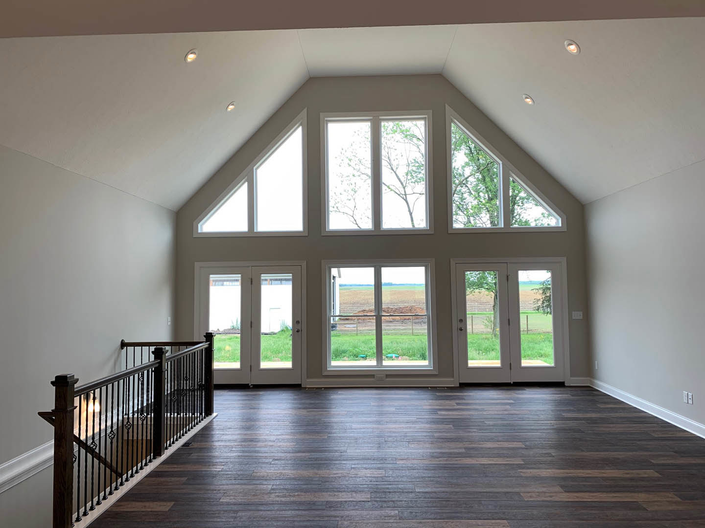 Spacious room featuring wood flooring, black metal staircase railing, multiple large windows with views of trees and open field, natural daylight illuminating interior.