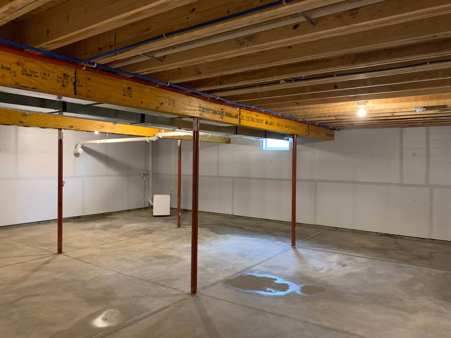 Concrete floor with puddle of water and blue spot, exposed wooden beam and pipes on ceiling, white box on floor, unfinished walls with building insulation