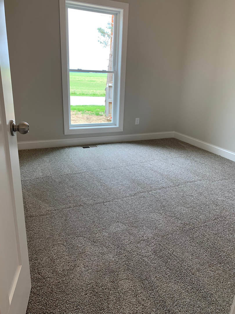 Carpeted room with white walls, large window overlooking grassy field, white pillar, door with silver knob, floor vent