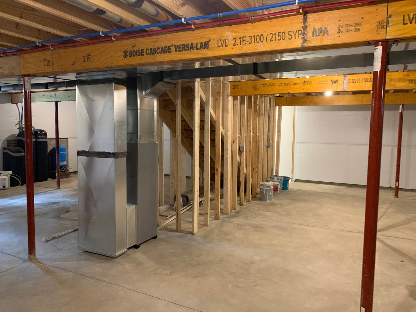 Open room with exposed steel ceiling beams, concrete floor, refrigerator against a wall, grey paint bucket with white paint, yellow sign with black lettering hanging from ceiling