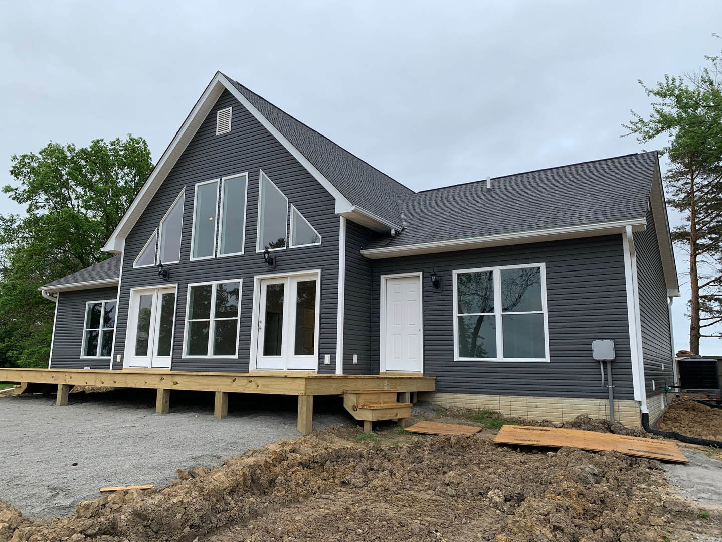 Modern home exterior with large windows reflecting surrounding trees, wooden deck, metal siding, white door set in black brick wall, and dirt landscaping under mature trees