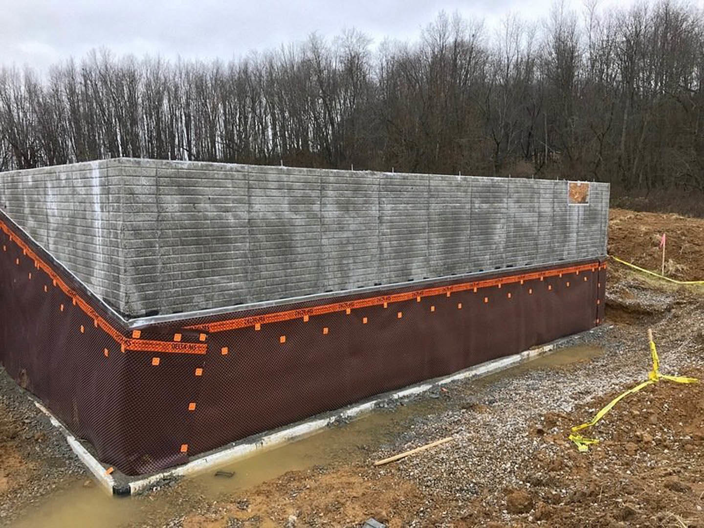 Concrete wall with textured brown surface, orange and black construction tape, yellow tape on the ground, distant trees, grey brick wall illuminated by orange light