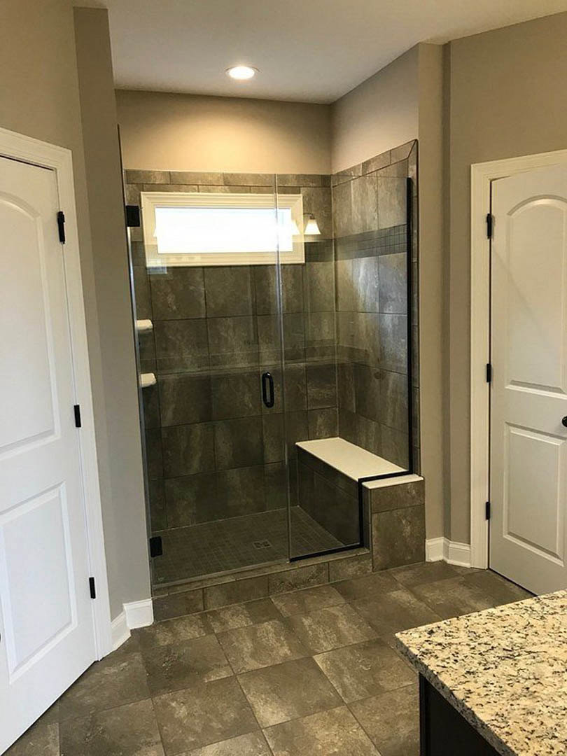 Glass shower enclosure with built-in bench, white door featuring black handle, white framed accent on exposed brick wall, close-up of tiled floor and countertop, cabinetry and