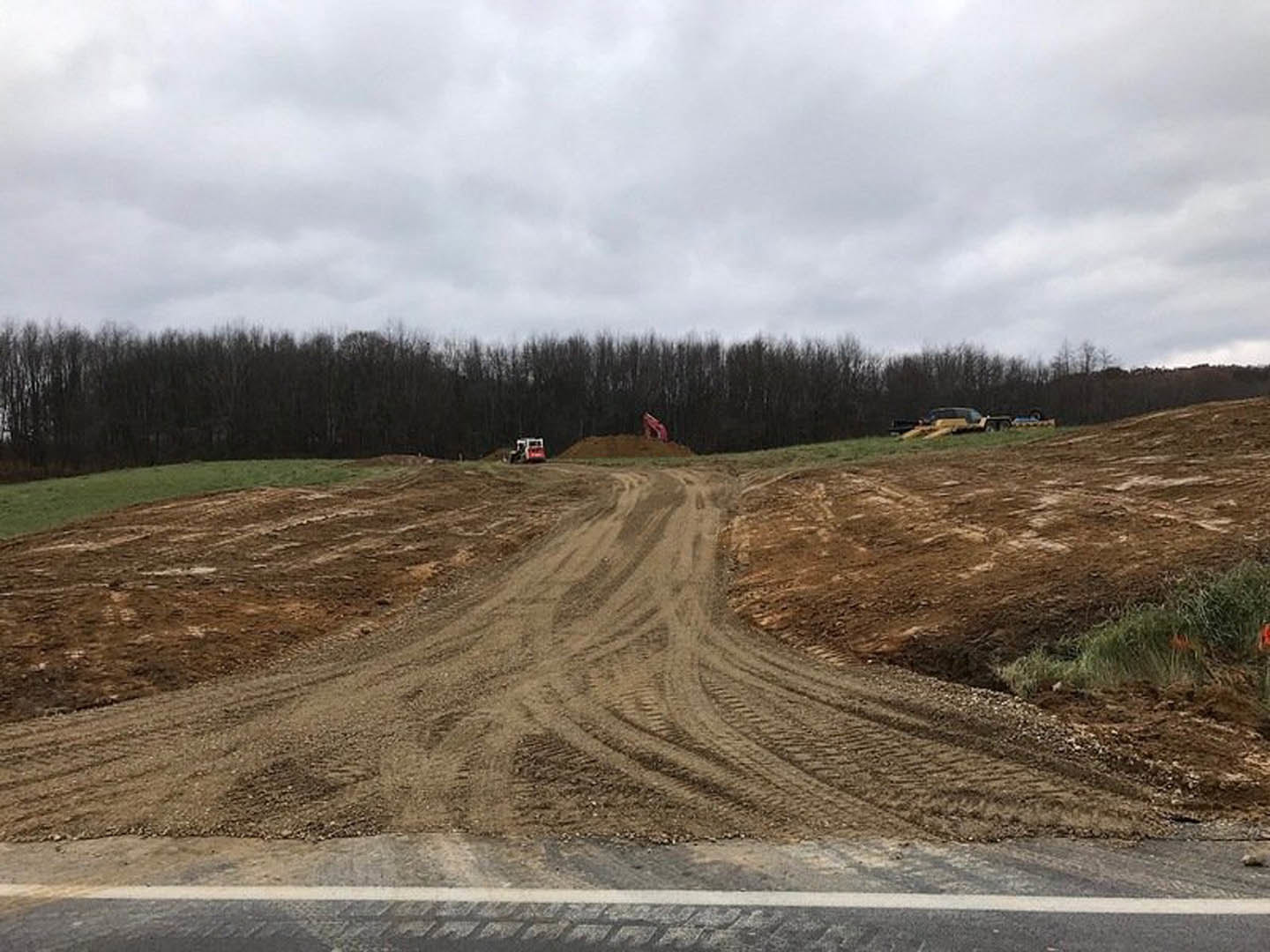 Dirt road with tire tracks leading to a dirt pit, surrounded by grass and dense trees under a cloudy sky