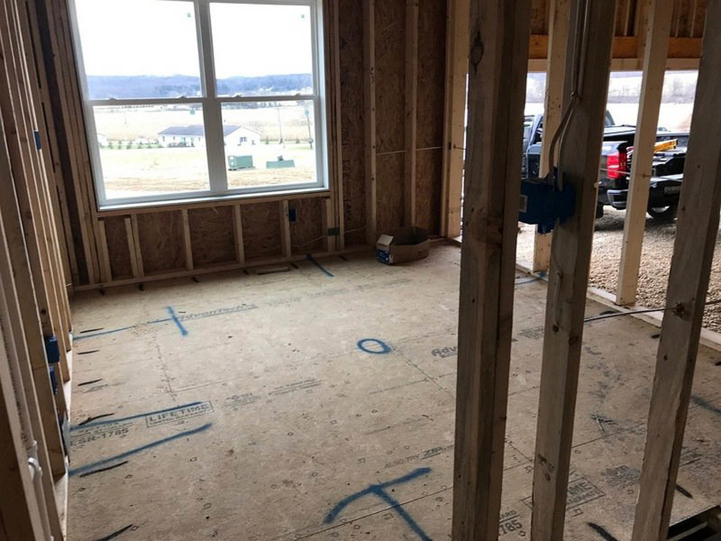 Unfinished interior with exposed wooden floors, large window overlooking grassy field, construction materials scattered, parked truck visible outside