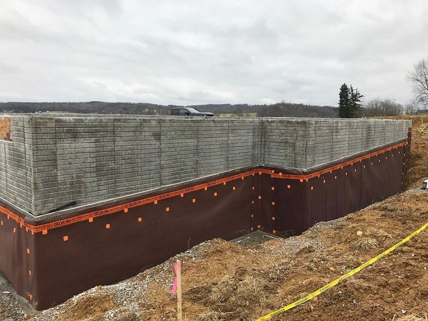 Brick wall under construction with yellow caution tape on dirt, leafless tree, parked car, and cloudy sky in background