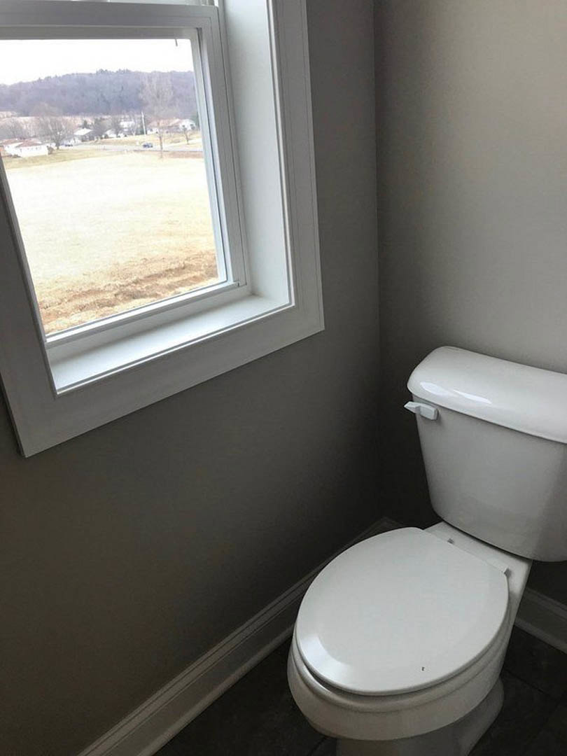 White porcelain toilet with lid up and seat down, set against light-colored bathroom wall, adjacent to window and plumbing fixtures.