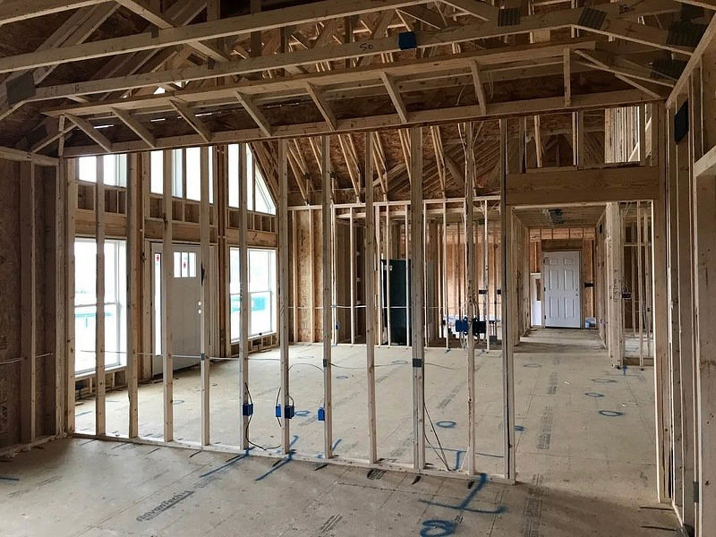 Wood-framed room under construction with exposed ceiling beams, white-framed window, white door with black frame, and sunlight casting a person’s shadow on the unfinished floor.