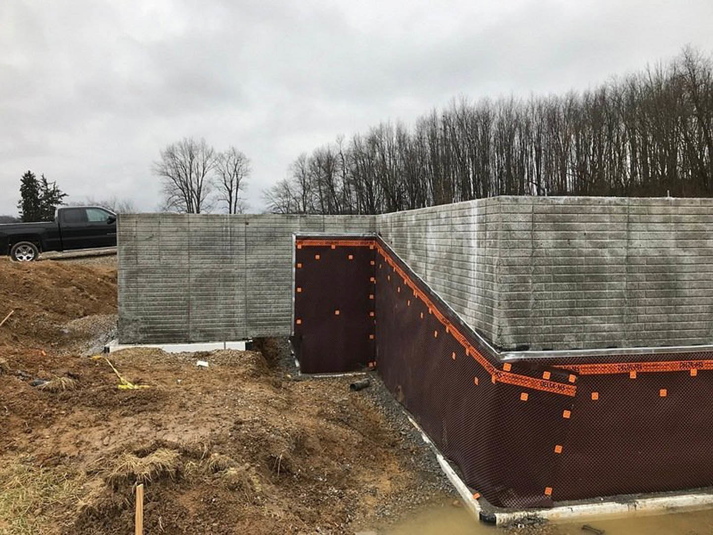 Partially built home with exposed brick wall, orange and black plastic sheeting, black truck parked behind, and trees visible in the background