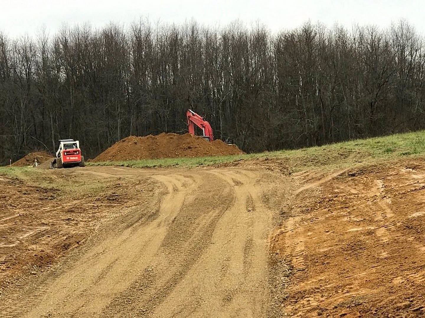 Dirt road bordered by grass and leafless trees, red excavator parked beside a large pile of soil under an open sky
