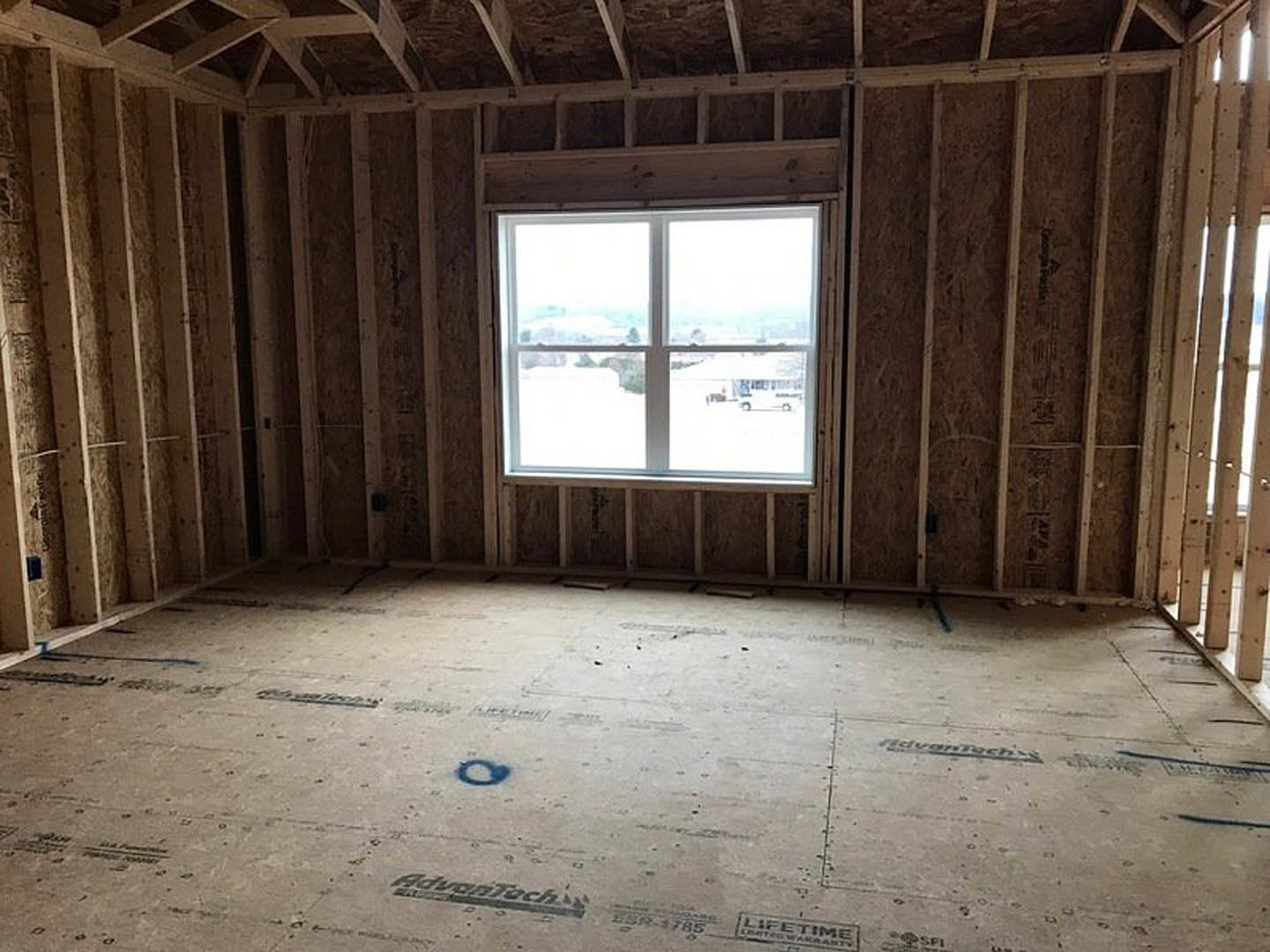 Sunlit room with wide window, natural wood plank flooring, exposed wooden ceiling beam, blue accent paint along floor trim, white walls, view of parking lot through window.