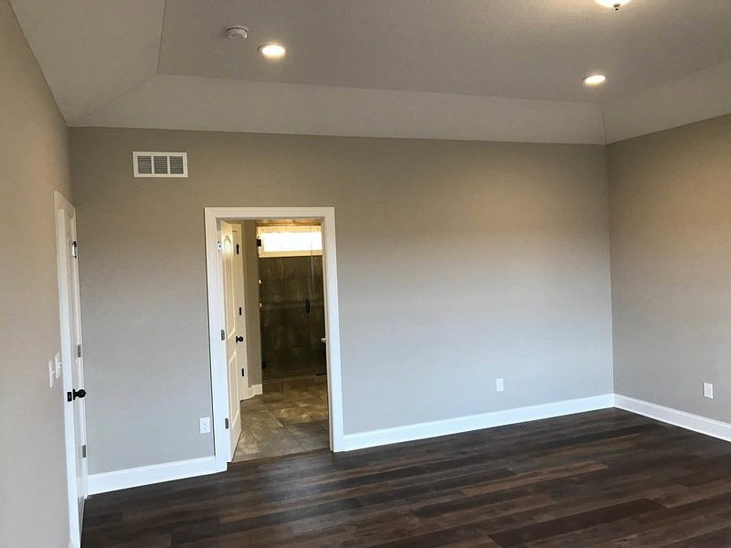 Dark wood flooring with white baseboard trim, open white door leading to adjacent room, ceiling light fixture, partial view of wall vent, and glimpse of tiled shower enclosure.