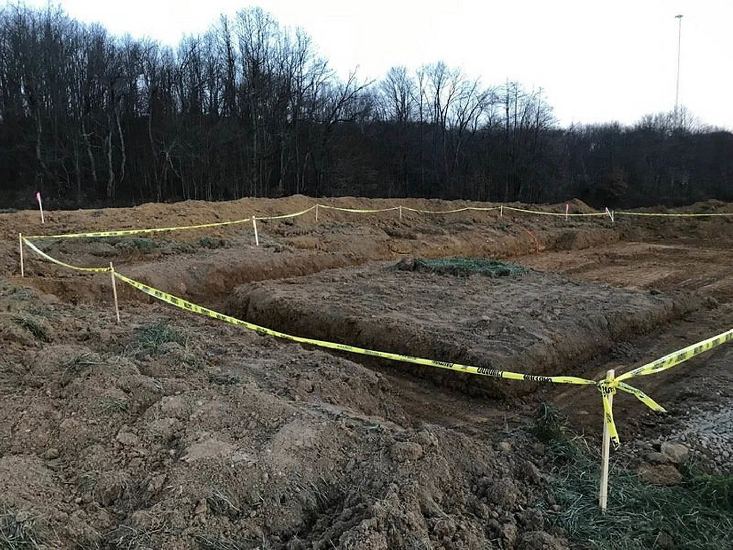 Dirt field marked with yellow tape, surrounded by trees and patches of grass under open sky