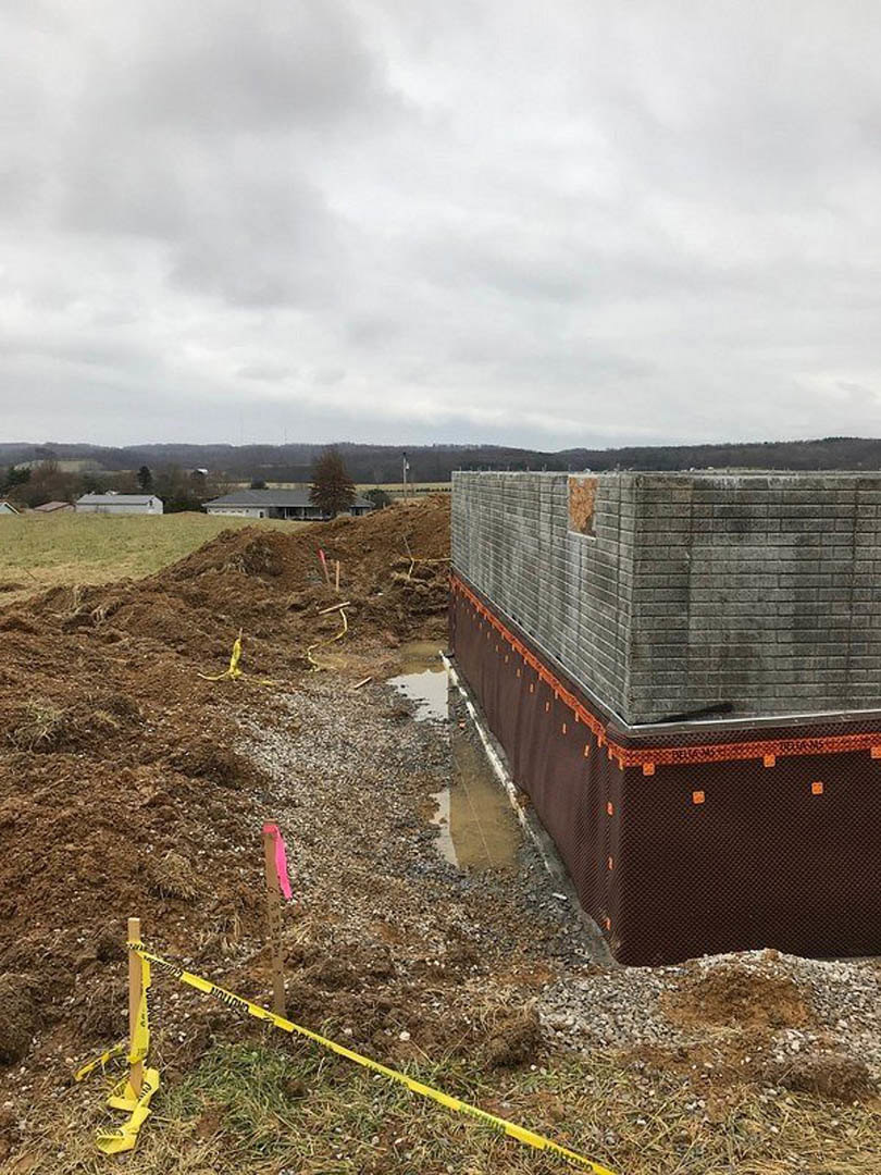 Partially built brick wall surrounded by brown protective cloth, leafy tree with yellow tape, grassy construction site under cloudy sky