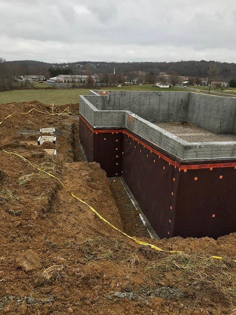 Concrete foundation with partial brick wall on a construction site, yellow caution tape in dirt, cloudy sky and grassy field in background
