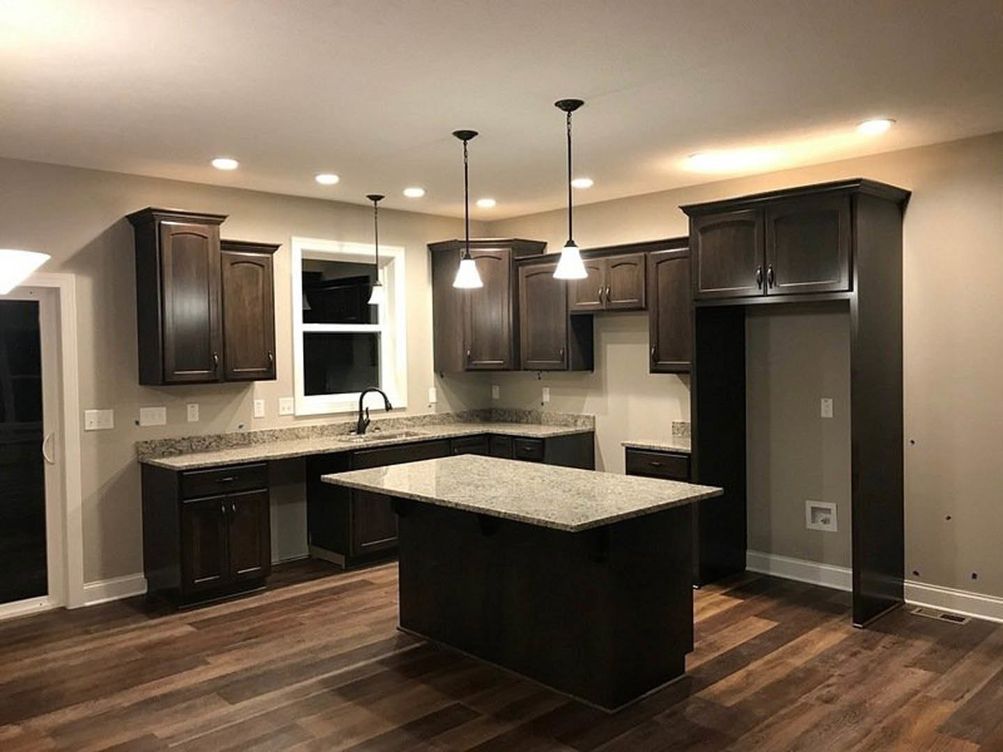 Dark wood kitchen cabinets, marble-topped island, stainless steel faucet, tile backsplash, large window with natural light, light-colored flooring