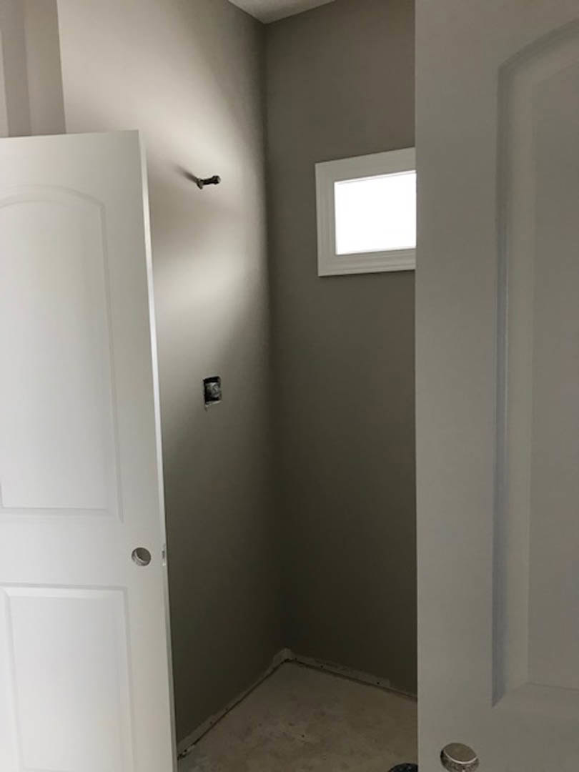 White paneled door with brushed metal handle set in a smooth white wall, corner of room visible with light wood flooring and recessed ceiling light.