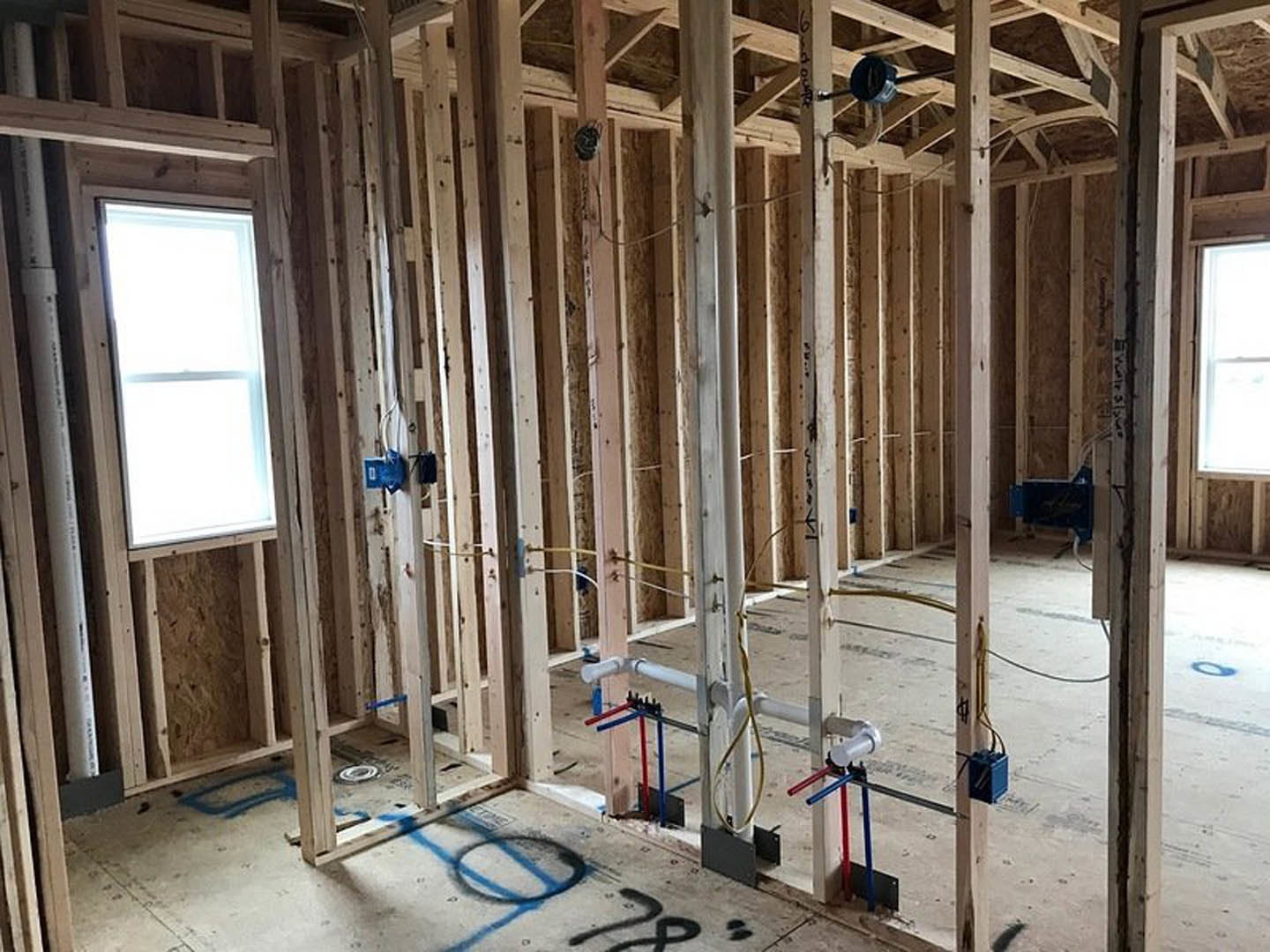 Exposed wood framing and beams with insulation, unfinished walls, visible plumbing pipes, ladder, and construction materials inside a custom home under construction