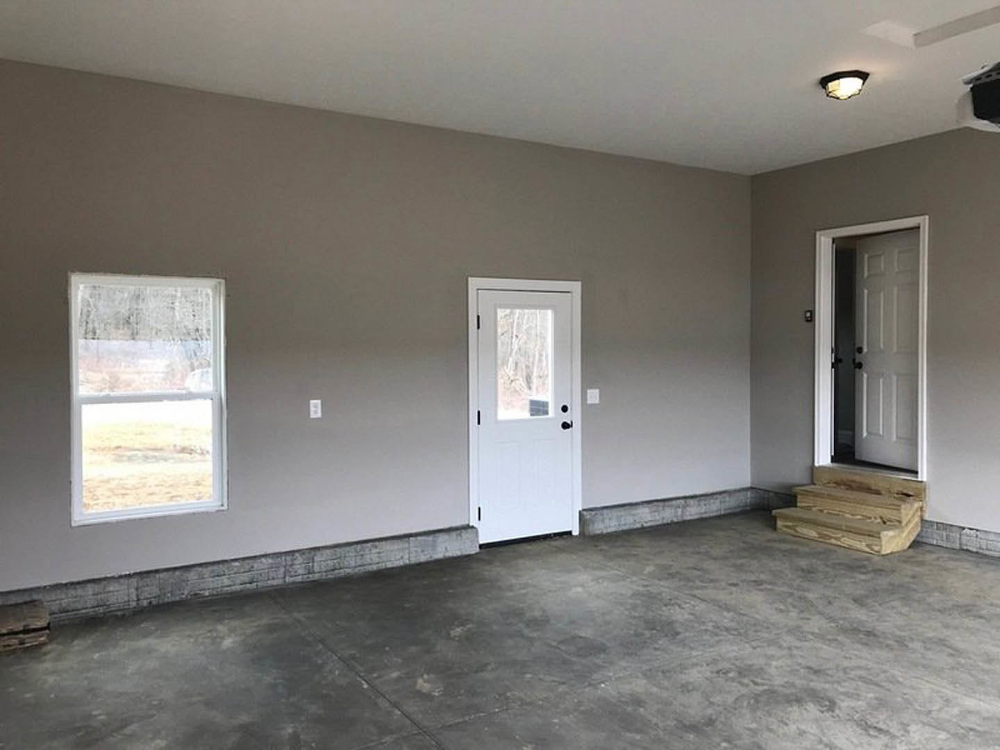 White paneled door with black hardware next to a wooden staircase, concrete floor, white walls, and window with white trim