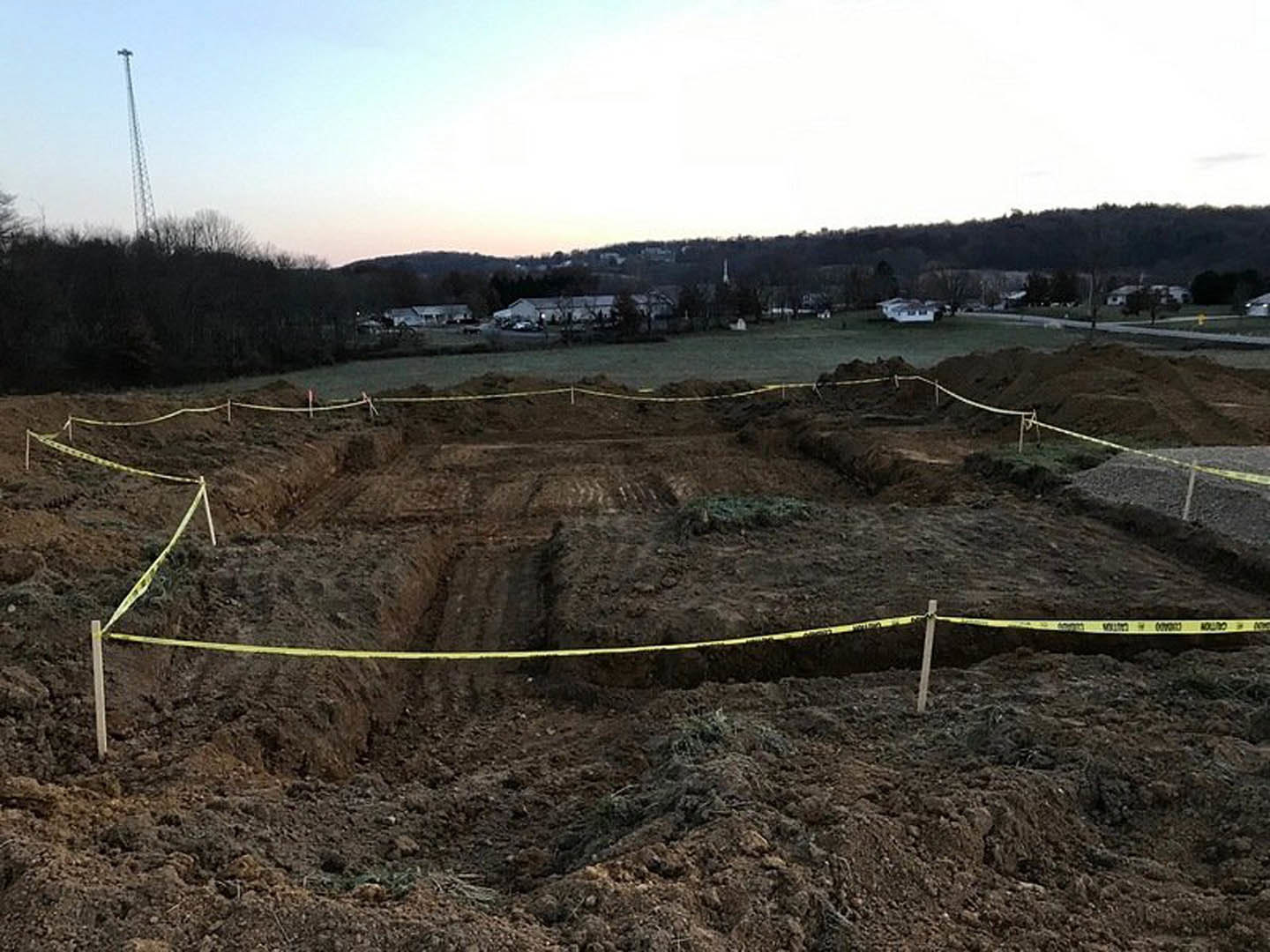 Construction site with yellow caution tape, dirt field, grassy patches, partially built structure, trees in background, blue sky with scattered clouds
