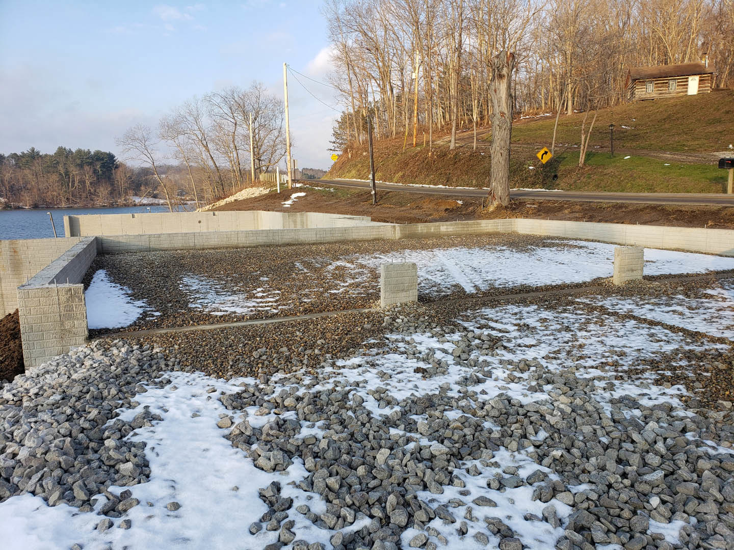 Log cabin with white door surrounded by snow-covered ground, pile of rocks, tree trunks, concrete block, and power lines on a hill under cloudy winter sky.