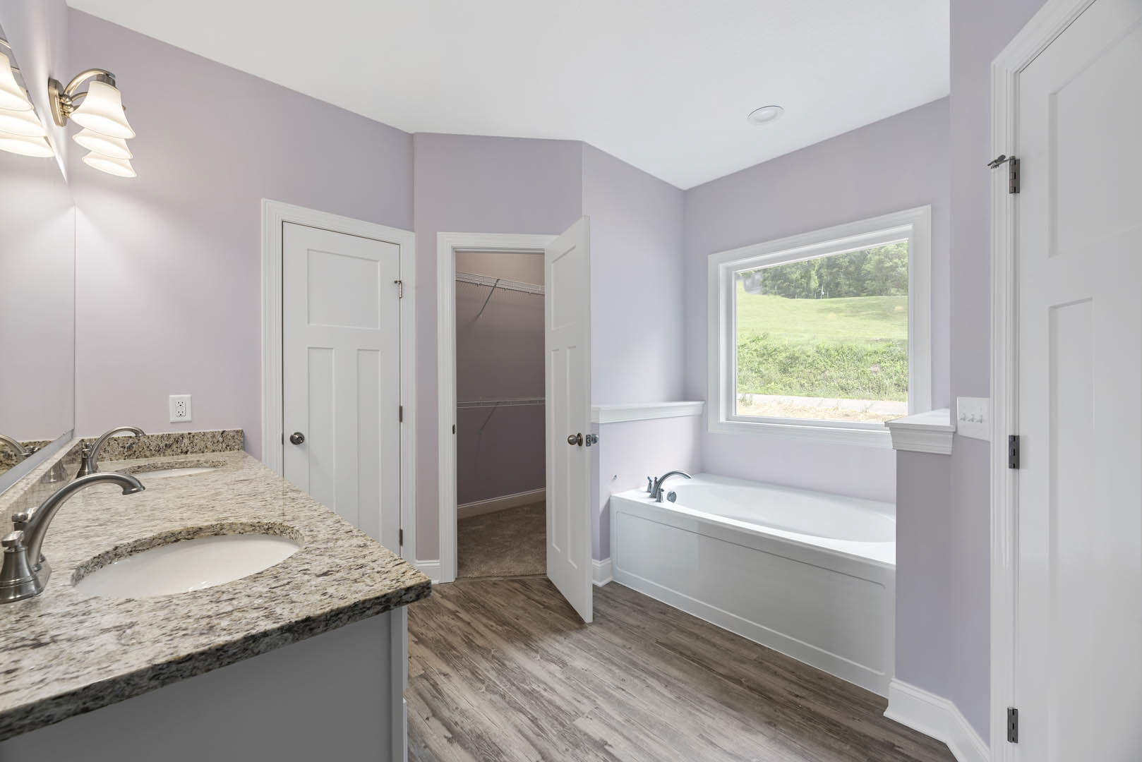 Freestanding bathtub beside a large window overlooking a field, white ceramic sink with chrome faucet on a stone countertop, light fixture mounted above, neutral tile walls and
