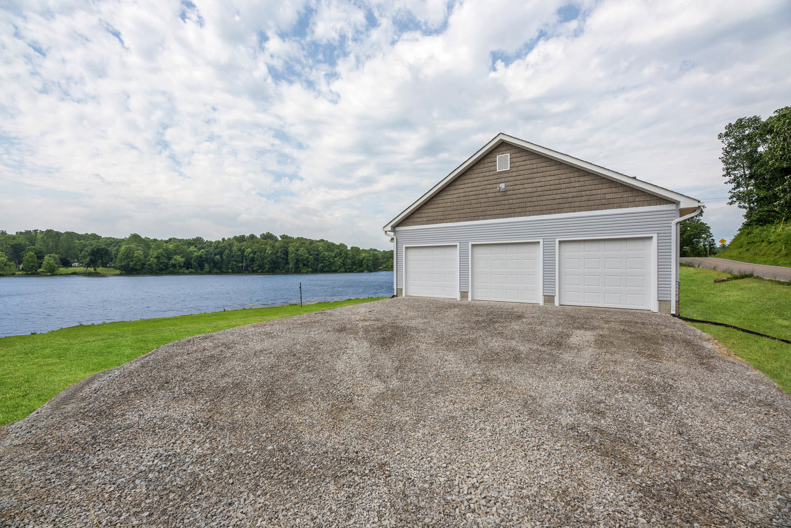 Three-door white garage with gravel driveway, surrounded by grass and trees, lake visible in the background under a partly cloudy sky