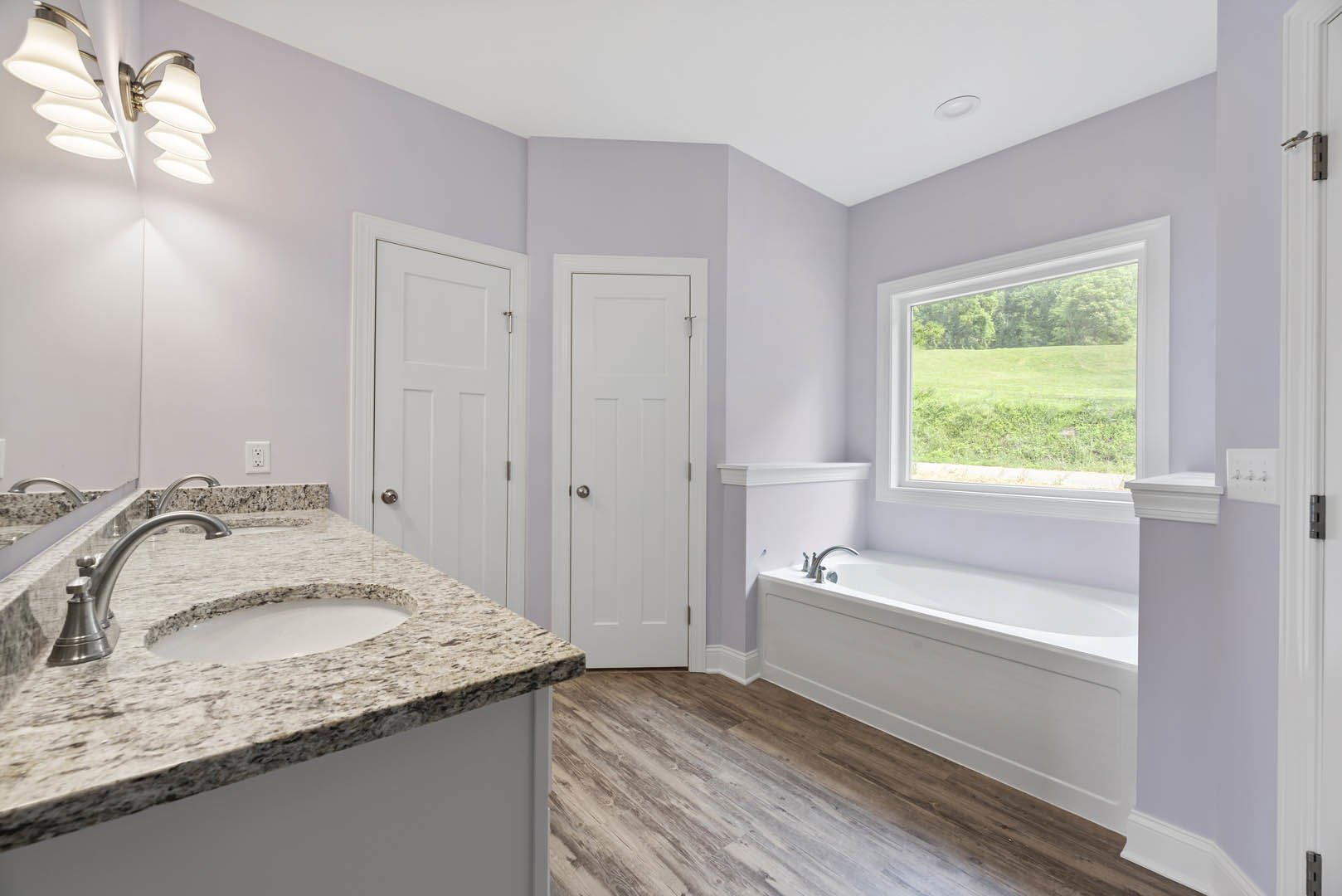Freestanding white bathtub and rectangular sink on stone vanity, large window overlooking grassy field, white paneled door with silver knob, modern chrome faucet, glass globe light