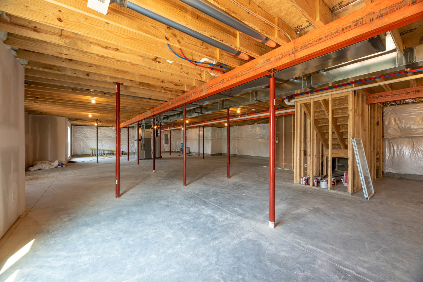 Room with exposed red metal poles, wooden plank ceiling, concrete floor, white rectangular vent with holes, pipes mounted on wood wall, and a white plastic bag resting on a bed.