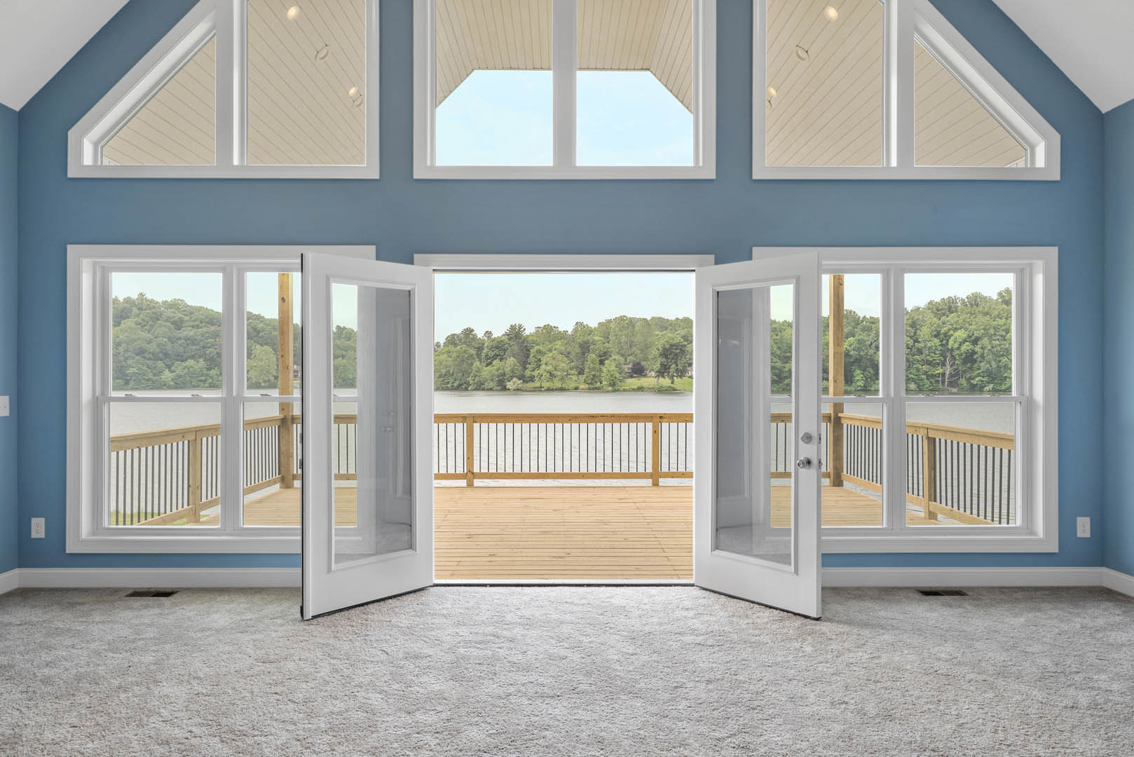 Open living area with light wood flooring, sliding glass doors leading to a deck, wooden railing, and expansive water view framed by forest through large windows