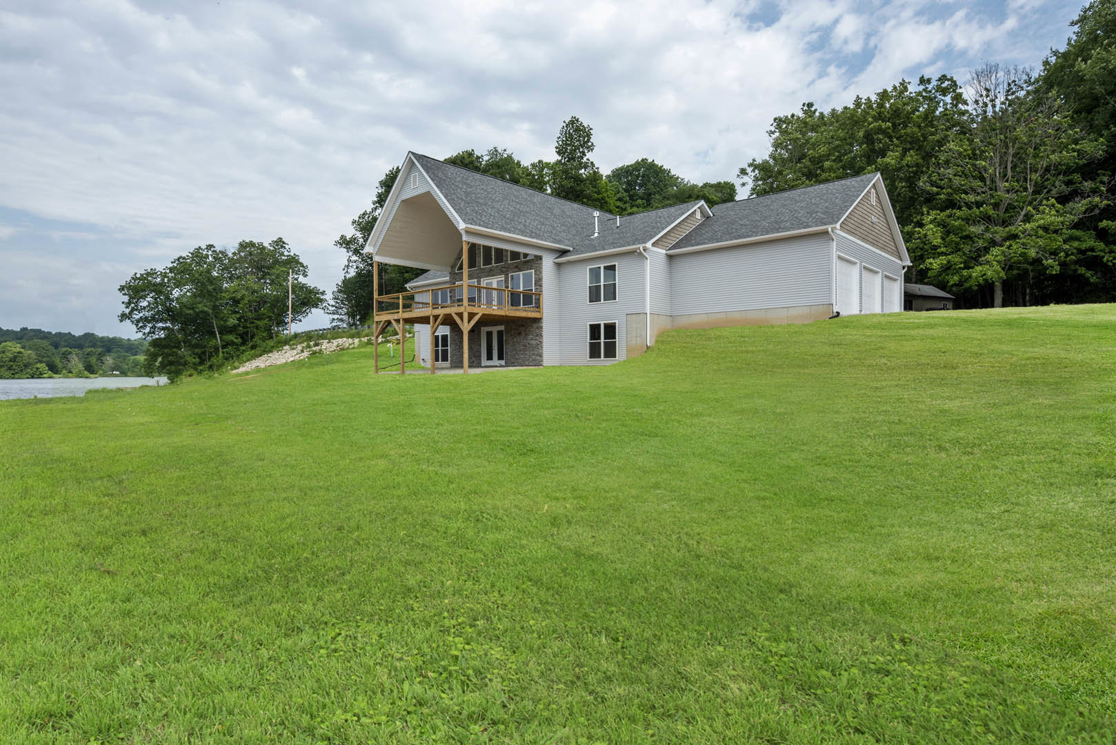 White farmhouse with wood side deck, expansive green lawn, and mature trees under partly cloudy sky
