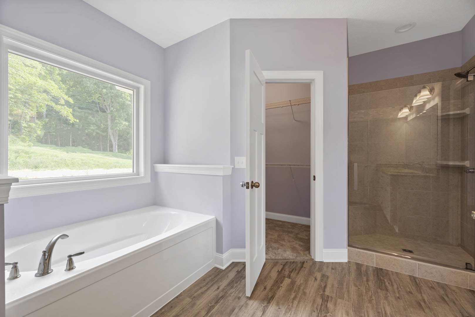 Modern bathroom with freestanding bathtub, glass-enclosed shower, large window overlooking trees, light tile flooring, white walls, and open door leading to closet