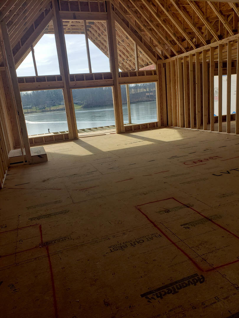 Open-concept room featuring large windows overlooking water and trees, wood beam ceiling, light-colored flooring with red lines, and modern finishes.