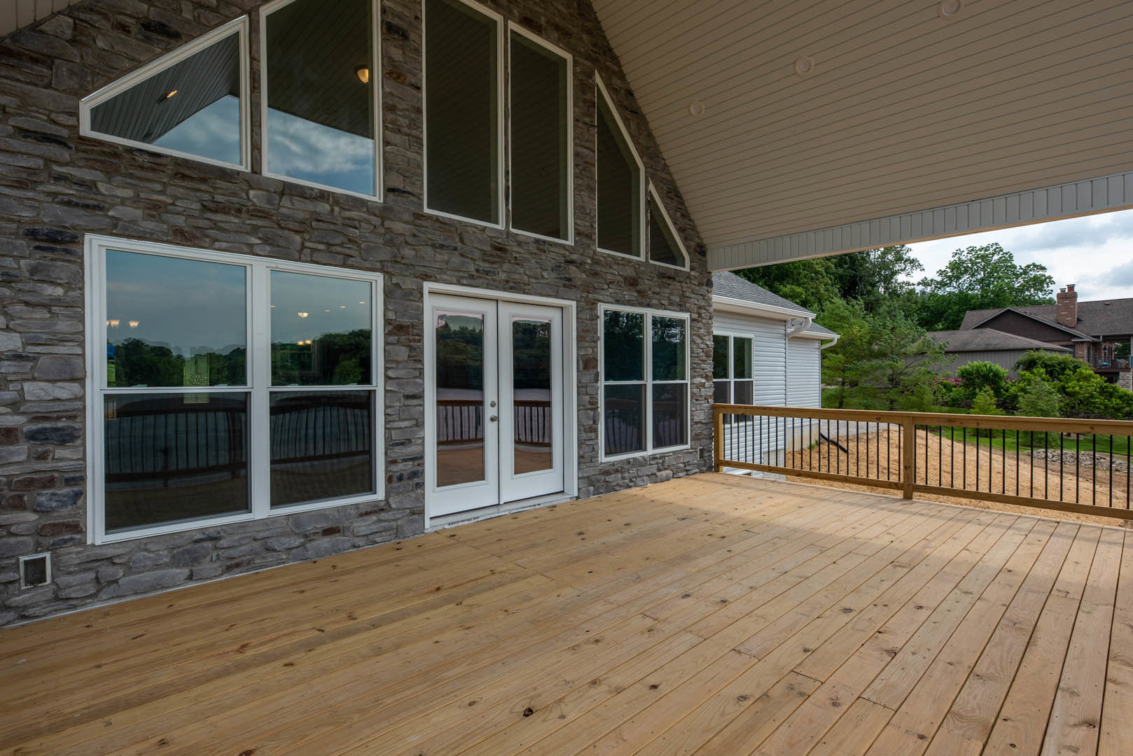 Large stone exterior home with wooden deck and railing, glass double doors, brick wall with window, and patio surrounded by stone walls.