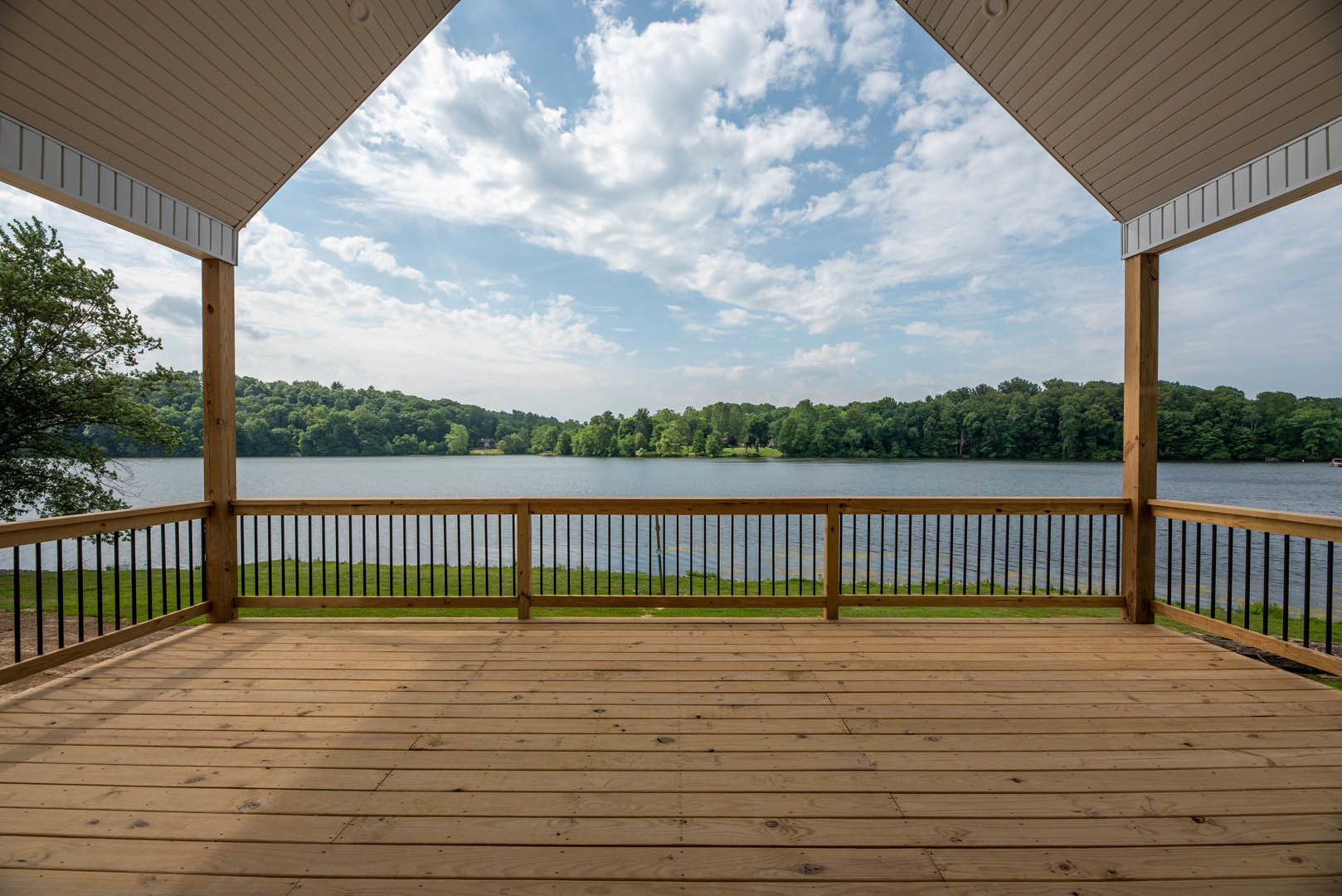 Wooden deck with metal railing, overlooking a lake and leafy trees under a blue sky with scattered clouds